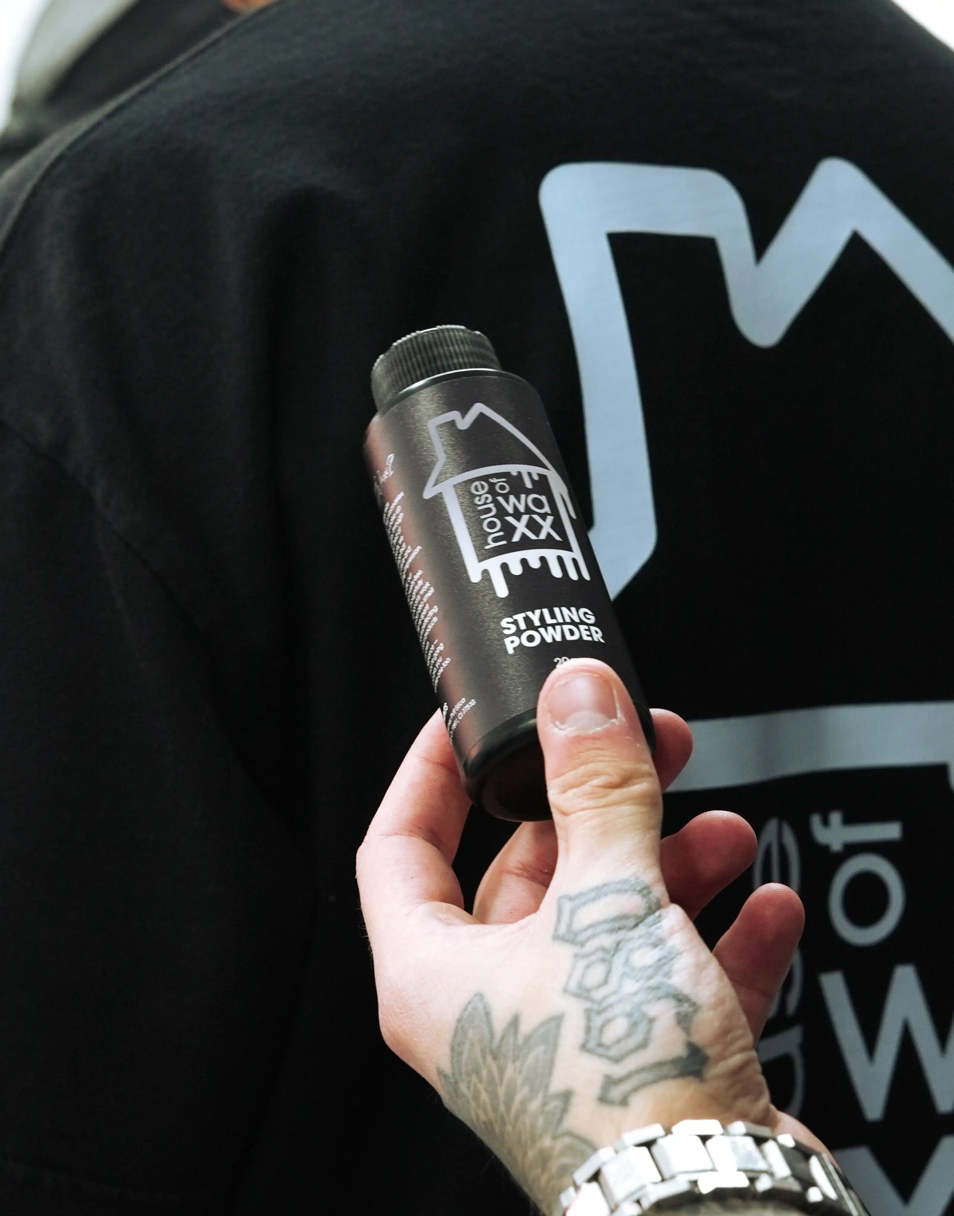 Tattooed hand holding a bottle of House of Waxx Styling Powder in front of a branded black t-shirt with the House of Waxx logo