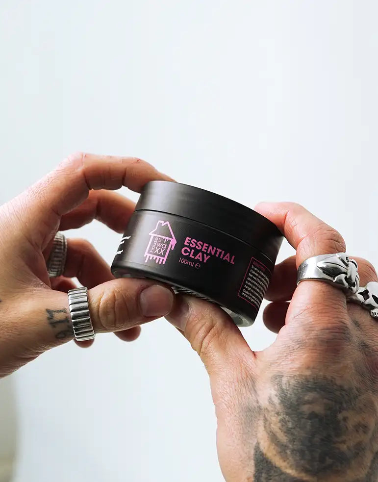 A tattooed mans hand holding a pot of House of Waxx Essential Clay 100ml on a white background
