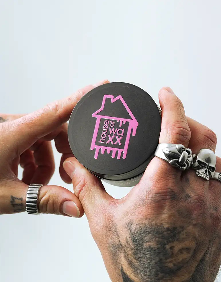 A tattooed mans hand holding a pot of House of Waxx Essential Clay 100ml on a white background