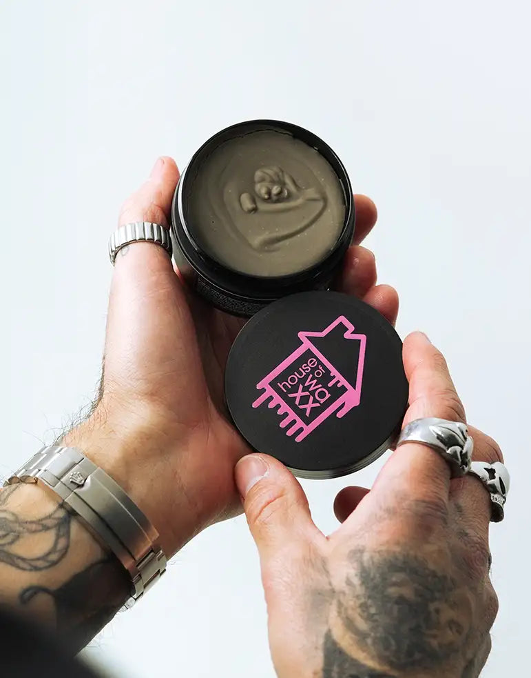 Tattooed hands holding an open jar of House of Waxx Essential Clay with a pink logo on the black lid, showcasing the product texture